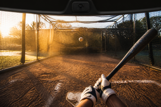 First-person view from the batter's box as a pitch crosses the plate inside a sunlit batting cage