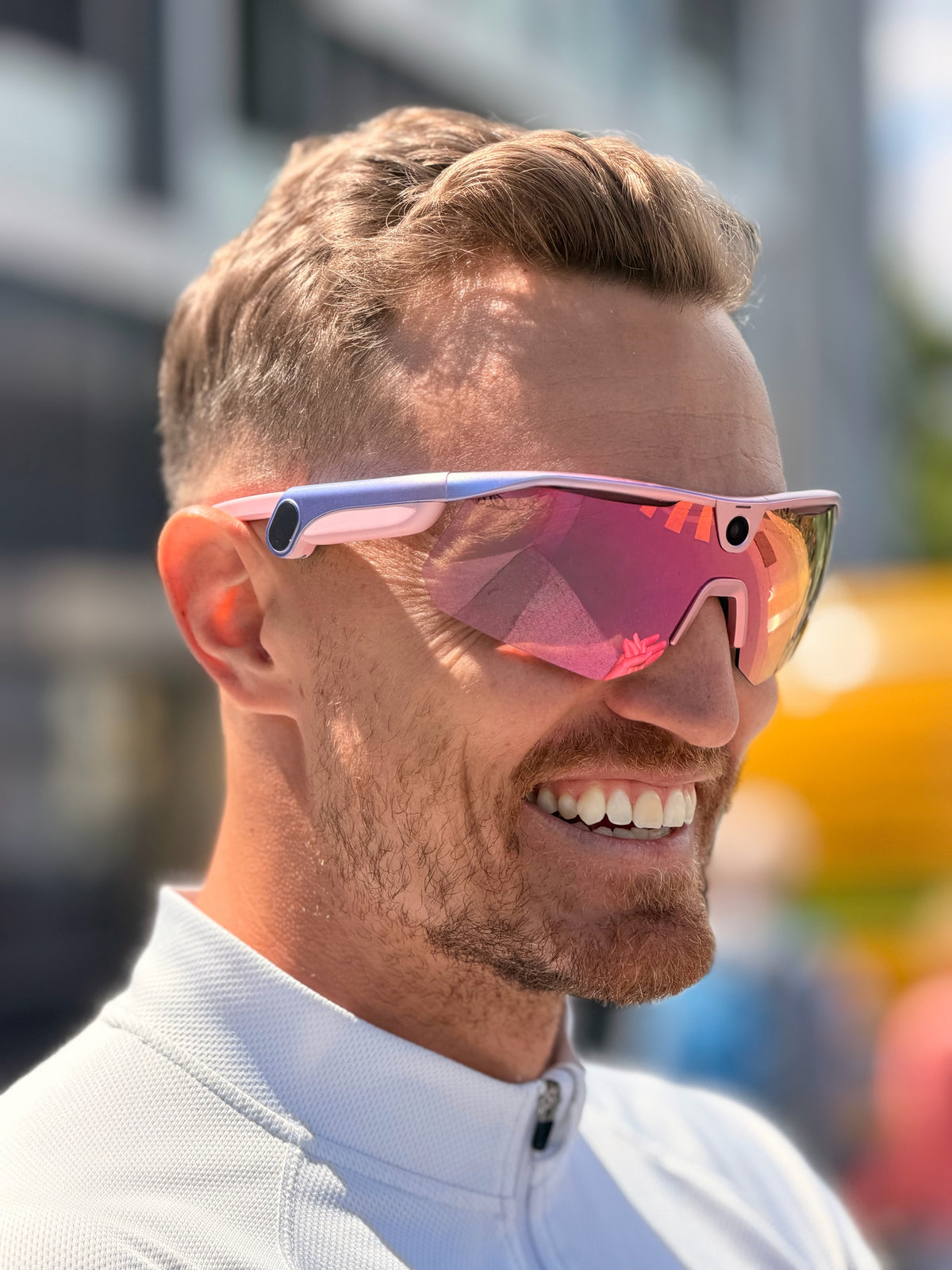 A man with short brown hair and beard smiling while wearing BLEEQUP Ranger AI Sports Camera Glasses with pink-framed sport sunglasses and mirrored lenses (UGC: @limitless.alex)